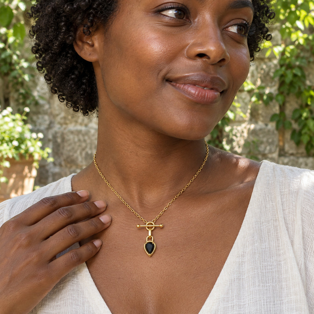 A woman in an ivory linen top wears the gold-tone black onyx necklace in a leafy courtyard with one hand beside her collarbone.