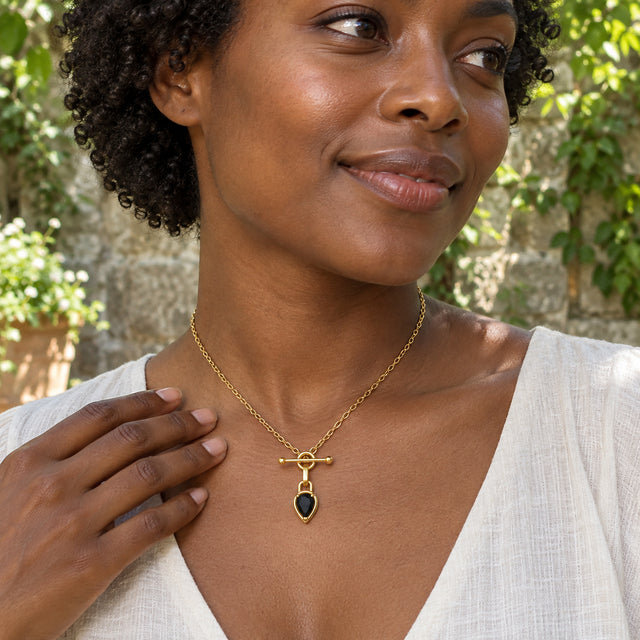A woman in an ivory linen top wears the gold-tone black onyx necklace in a leafy courtyard with one hand beside her collarbone.