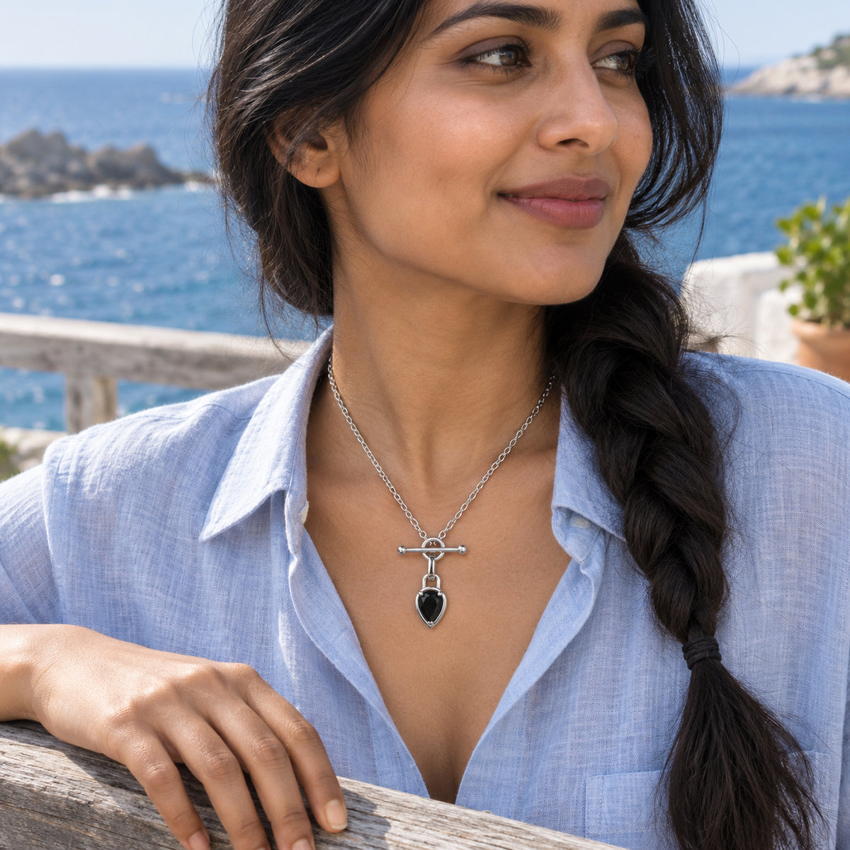 A woman with braided dark hair wears the silver-tone black pendant necklace with a pale blue shirt on a seaside terrace.