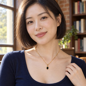 A woman in a navy top wears a gold-tone black onyx necklace beside a sunlit window.