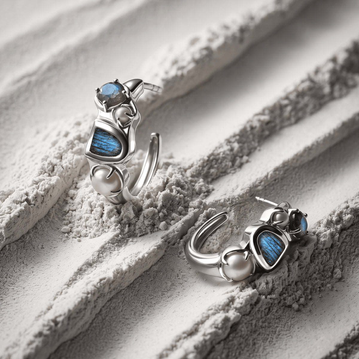 Two white rhodium labradorite and pearl hoops rest at different angles on a textured pale surface.