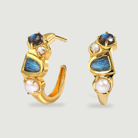 Two yellow gold plated hoops stand upright with blue labradorite stones and white pearls on a pale gray surface.
