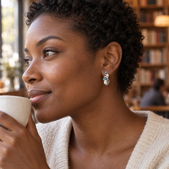 A woman with short curly hair wears a silver-colored labradorite and pearl hoop while holding a white ceramic cup in a cafe with bookshelves.