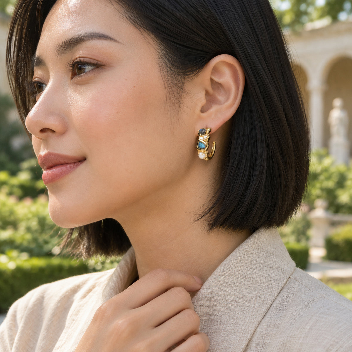 A woman with a straight black bob wears a yellow gold plated labradorite and pearl hoop beside garden greenery and pale stone arches.