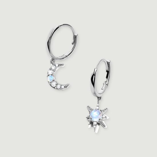 A pair of silver hoops with a crescent moon and a starburst charm rests on a light gray surface.