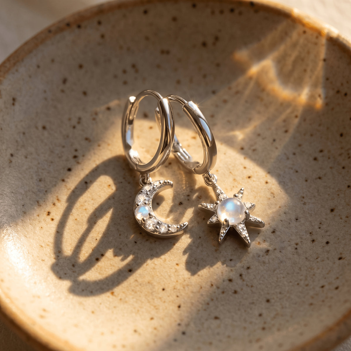 Silver crescent and starburst earrings sit in a speckled ceramic dish under warm window light.