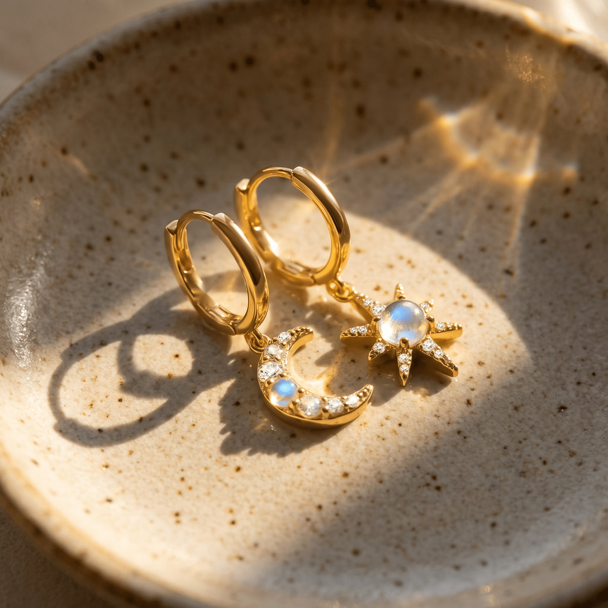 A yellow gold moonstone earring pair lies in a sunlit ceramic dish with curved shadows.