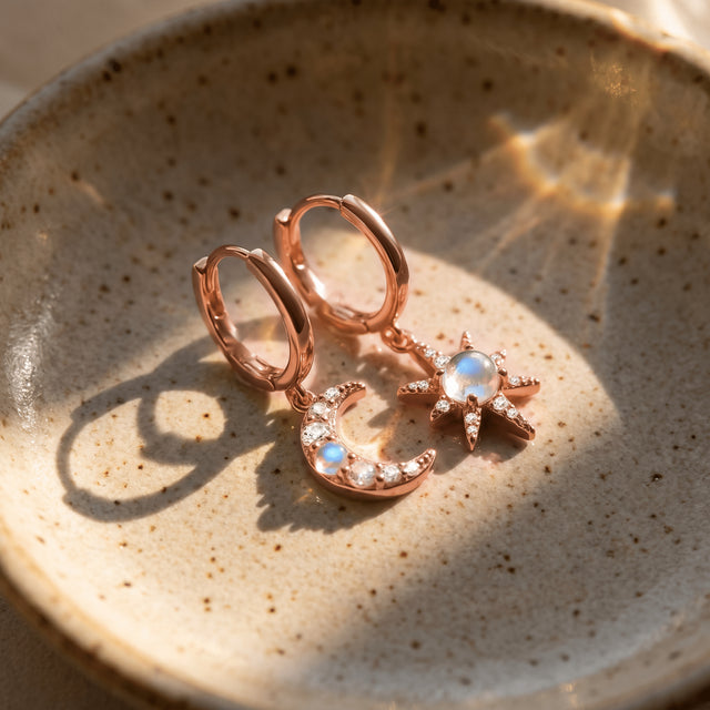 Rose gold crescent and starburst charms hang from hoops resting on a speckled ceramic surface.
