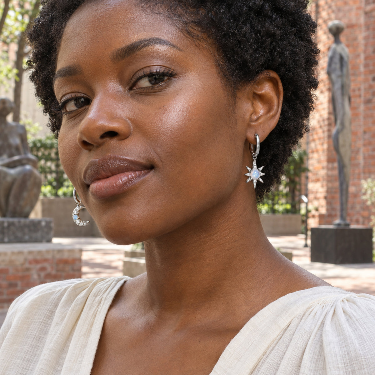 A woman with short natural curls wears silver crescent and starburst hoop earrings in a brick courtyard.