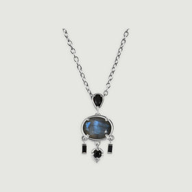A white rhodium plated necklace with an oval blue grey labradorite and black stone drops rests on a light grey background.