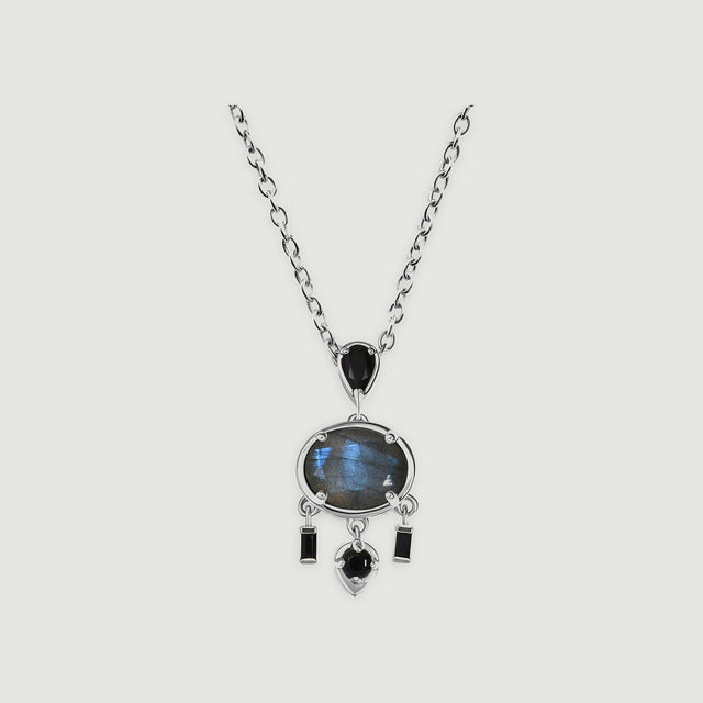 A white rhodium plated necklace with an oval blue grey labradorite and black stone drops rests on a light grey background.