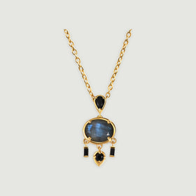 A yellow gold plated necklace displays an oval labradorite beneath a black pear shaped stone on a light grey surface.
