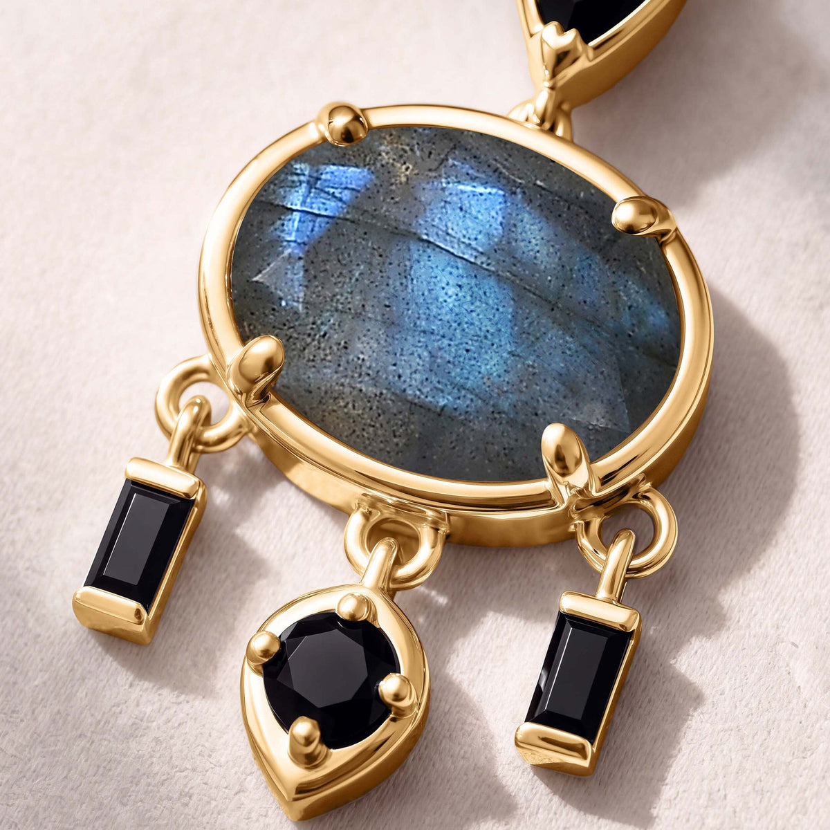 An oval labradorite with blue flashes sits in yellow gold plated prongs above two rectangular black stones and a round black drop.