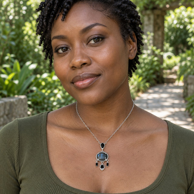 A woman with short twists wears a white rhodium plated labradorite necklace with an olive green top beside a garden path.