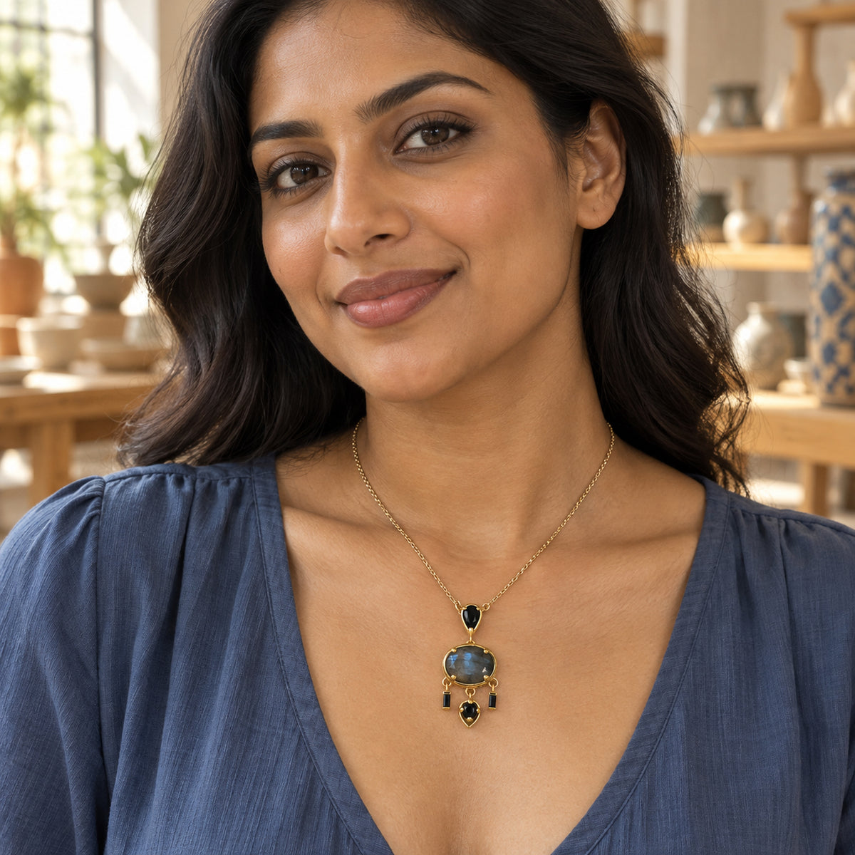 A woman with dark wavy hair wears a yellow gold plated labradorite pendant over a blue blouse in a pottery gallery.
