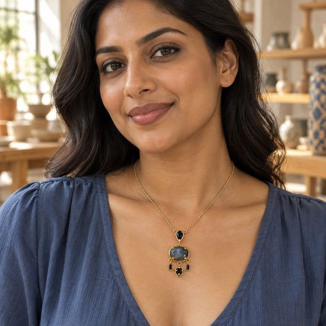 A woman with dark wavy hair wears a yellow gold plated labradorite pendant over a blue blouse in a pottery gallery.