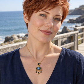 A woman with short copper hair wears a yellow gold plated labradorite necklace and a navy blouse beside the sea.