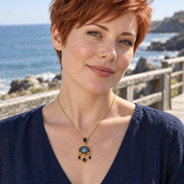 A woman with short copper hair wears a yellow gold plated labradorite necklace and a navy blouse beside the sea.