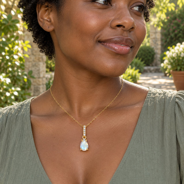 A woman in a sage green blouse wears a yellow gold moonstone necklace in a sunlit garden courtyard.