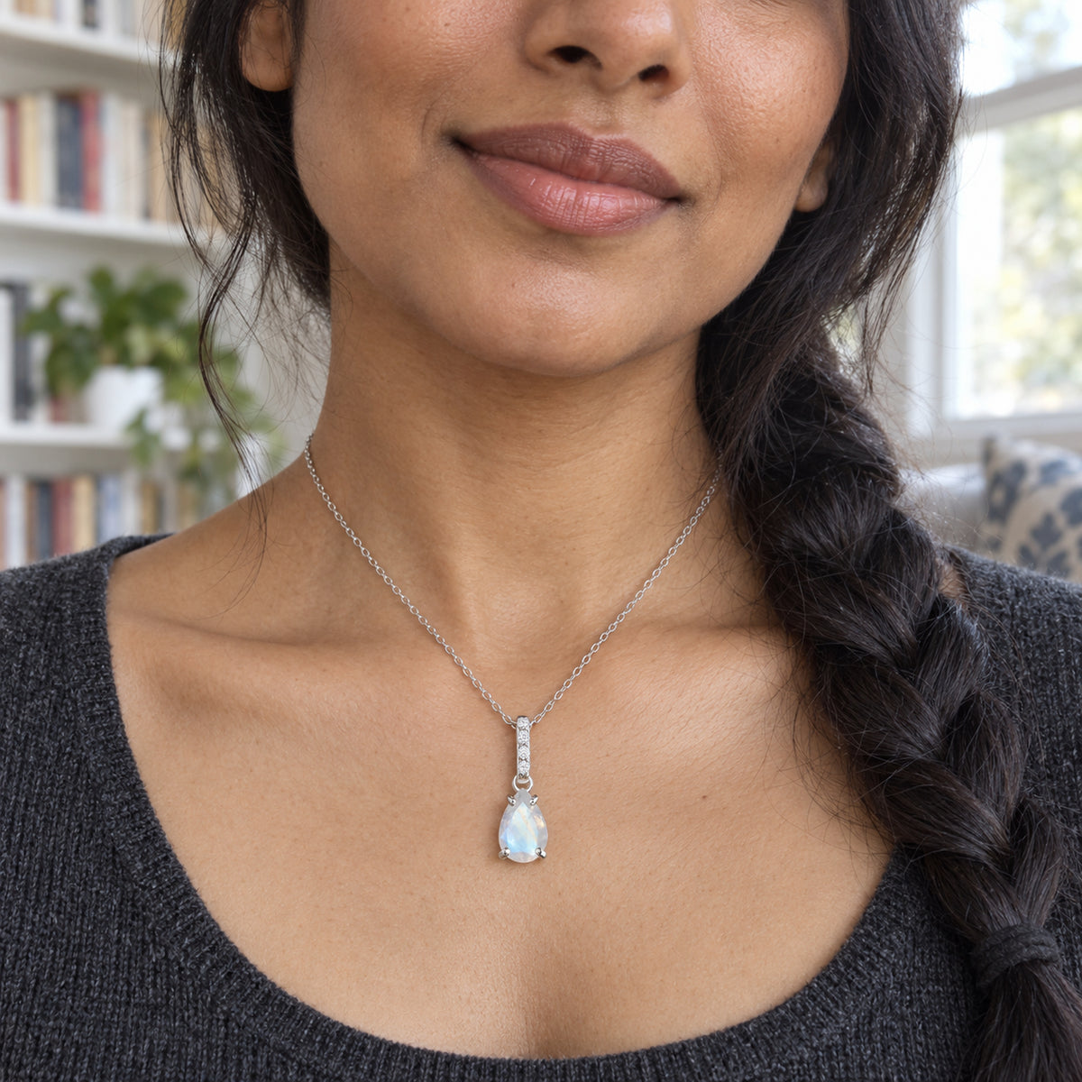 A woman with a dark braid wears a silver moonstone necklace above a charcoal knit neckline beside bookshelves.