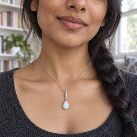 A woman with a dark braid wears a silver moonstone necklace above a charcoal knit neckline beside bookshelves.