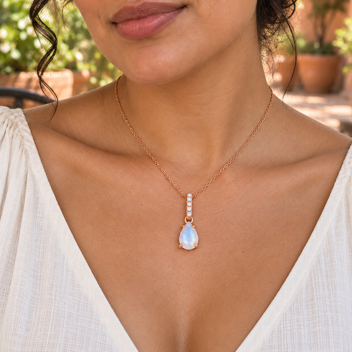A rose gold moonstone necklace rests against the chest of a woman in an ivory blouse on a leafy terracotta patio.
