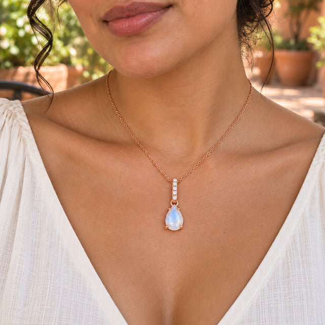 A rose gold moonstone necklace rests against the chest of a woman in an ivory blouse on a leafy terracotta patio.
