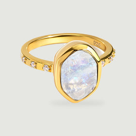 A yellow gold ring with an irregular pale moonstone and small clear stones rests on a light grey surface.