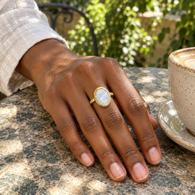A yellow gold moonstone ring is worn on a hand resting on a stone cafe table beside a ceramic cup.