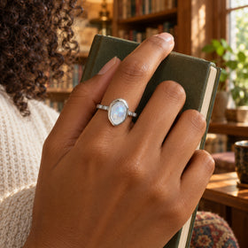 A silver moonstone ring sits on a finger as a hand holds a closed green notebook in a sunlit library.
