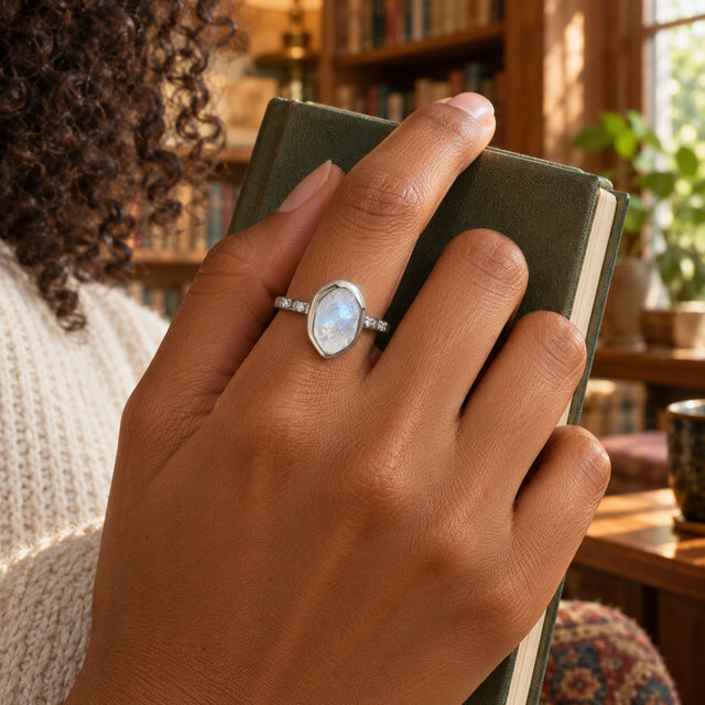 A silver moonstone ring sits on a finger as a hand holds a closed green notebook in a sunlit library.