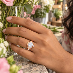 A rose gold moonstone ring is worn beside a glass vase of pink and white flowers on a stone counter.