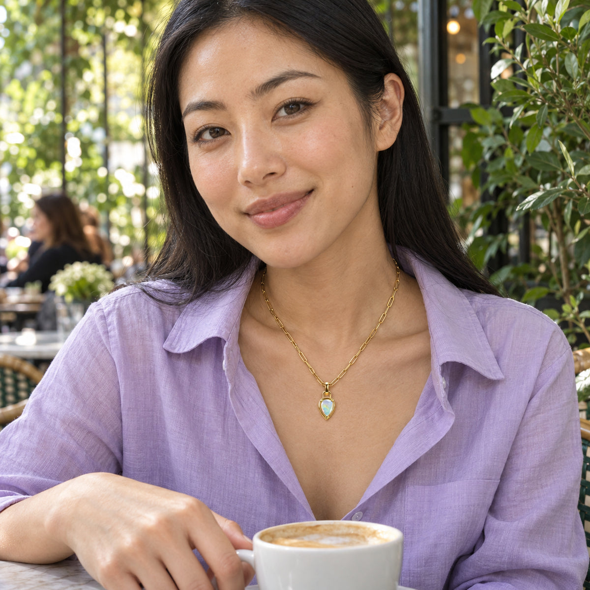 A woman in a lilac blouse wears the yellow gold opal necklace at a leafy cafe with a cup of coffee.