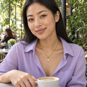 A woman in a lilac blouse wears the yellow gold opal necklace at a leafy cafe with a cup of coffee.