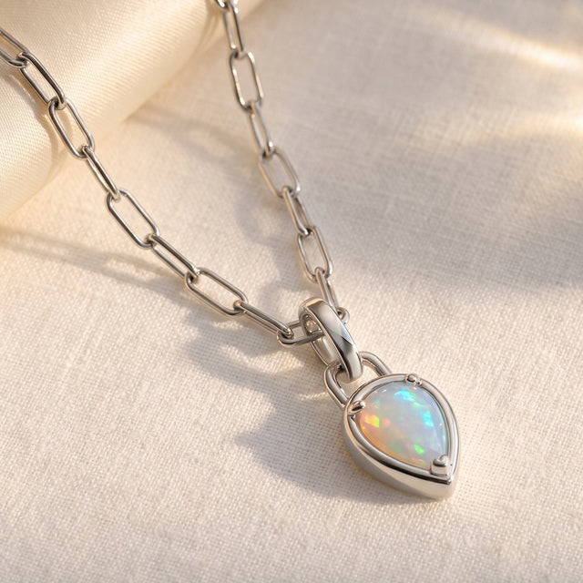 A silver-tone wide link chain leads to an opal pendant resting on softly folded ivory fabric.