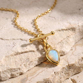 The yellow gold chain and opal pendant lie diagonally across a pale textured stone surface.