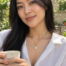 A woman with long black hair wears the yellow gold opal necklace with a white linen blouse while holding a ceramic cup in a leafy courtyard.