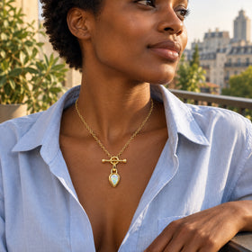 A woman with short curls wears the yellow gold opal pendant above an open blue shirt on a sunlit balcony.