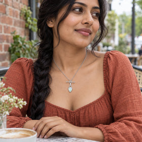 A woman with a dark braid wears the silver-colored opal pendant and a terracotta blouse at an outdoor cafe table.