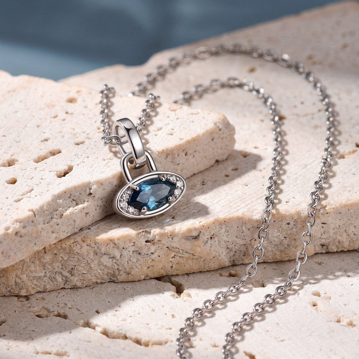 A white rhodium plated blue topaz necklace lies across pale textured stone with its chain curving beside the charm.