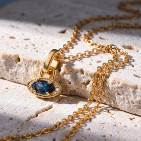 A yellow gold plated blue topaz charm rests on a pale stone ledge with the linked chain draped around it.