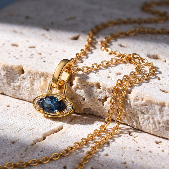 A yellow gold plated blue topaz charm rests on a pale stone ledge with the linked chain draped around it.