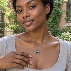 A woman in a gray scoop-neck top wears the white rhodium plated blue topaz necklace in a leafy brick courtyard.