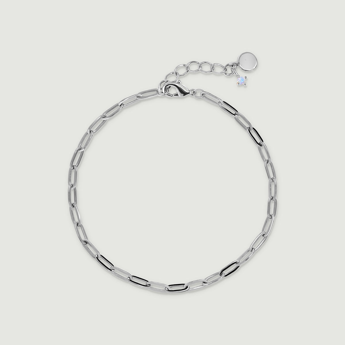 A white rhodium plated anklet with elongated open links and a small pale charm lies on light gray.