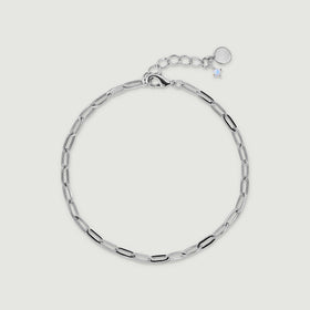 A white rhodium plated anklet with elongated open links and a small pale charm lies on light gray.