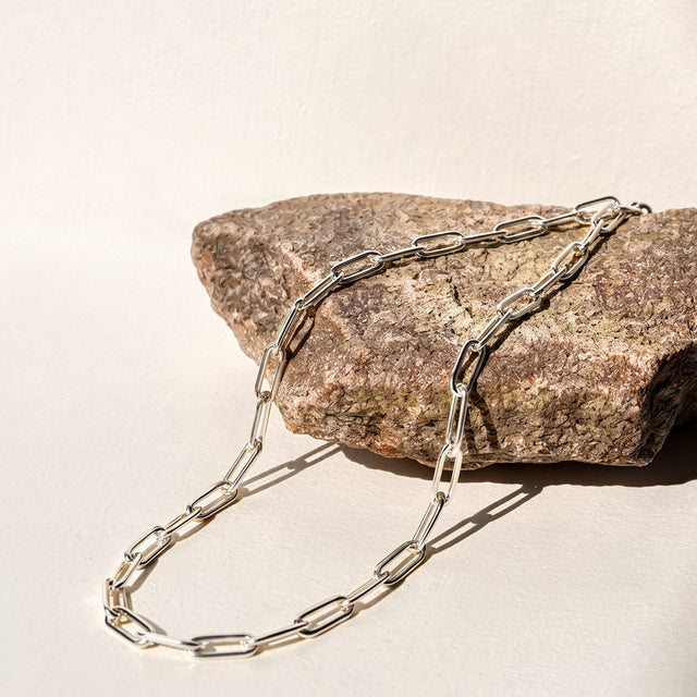 A white rhodium plated open-link anklet drapes over a textured stone on a pale tabletop.