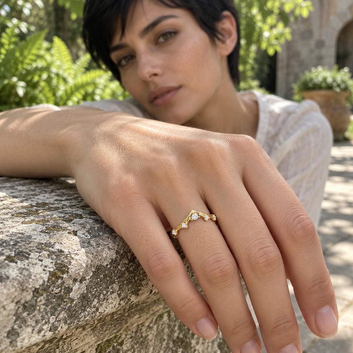 A yellow gold diamond ring is worn on a hand resting on a weathered stone ledge in a leafy garden.