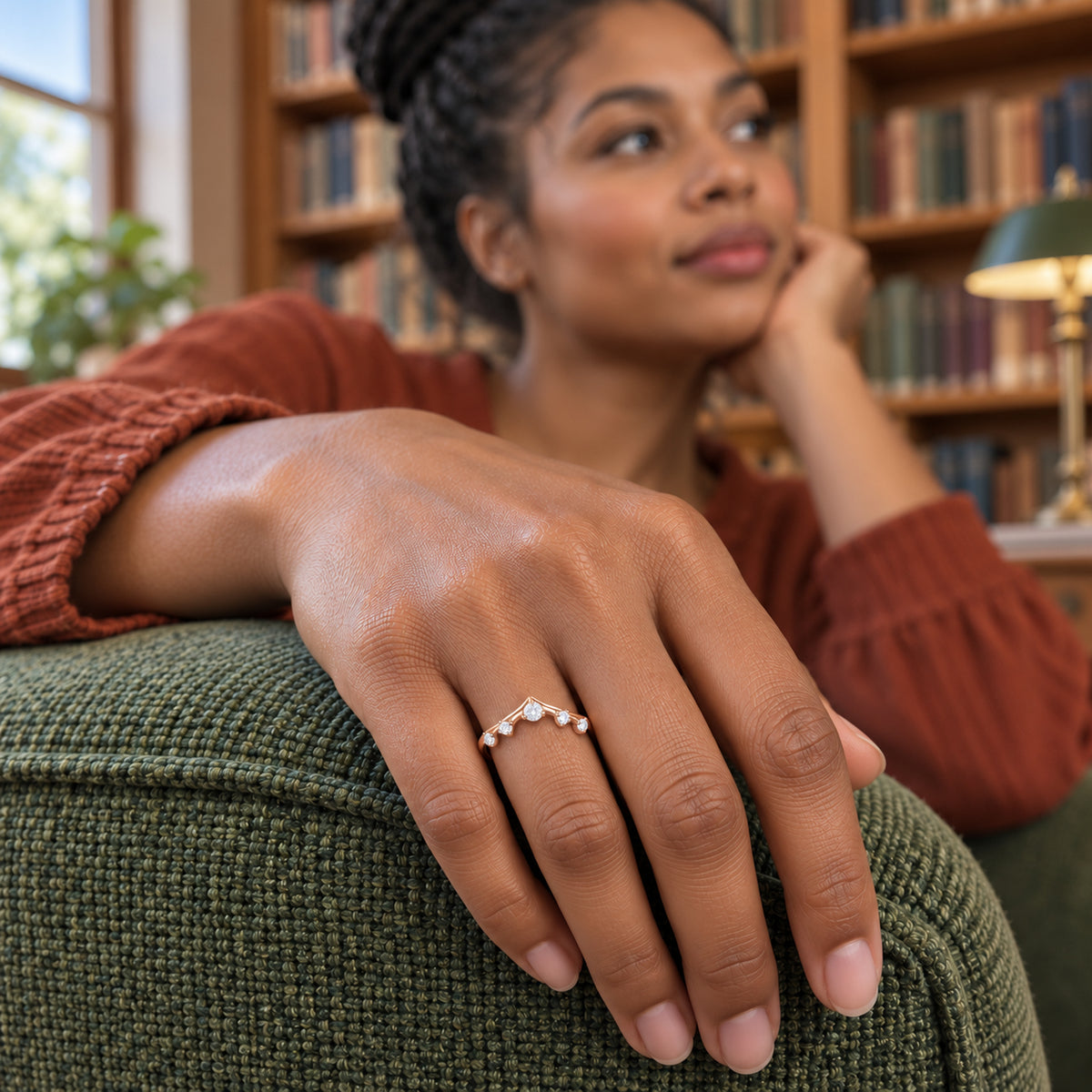 A rose gold ring with five round diamonds is shown on a hand resting on a green upholstered chair beside bookshelves.