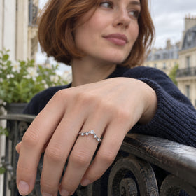 A white gold diamond ring is worn on a hand resting on a balcony railing, with a navy sleeve and city buildings behind it.