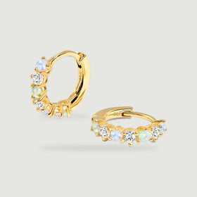 Two yellow gold hoops with pale blue, green, and clear round stones rest on a light gray background.